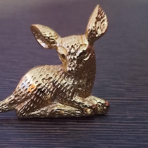 May include: A small, golden-colored figurine of a deer in a resting position. The deer has detailed fur texture and large, upright ears. The figurine is likely made of metal and sits on a dark, wood-grain surface.