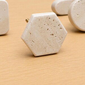 May include: A collection of cabinet knobs made from beige porous stone with natural pitting. The set includes a central hexagonal knob, a square knob to the left, and a circular knob to the right, each featuring a small brass-colored metal mounting post.