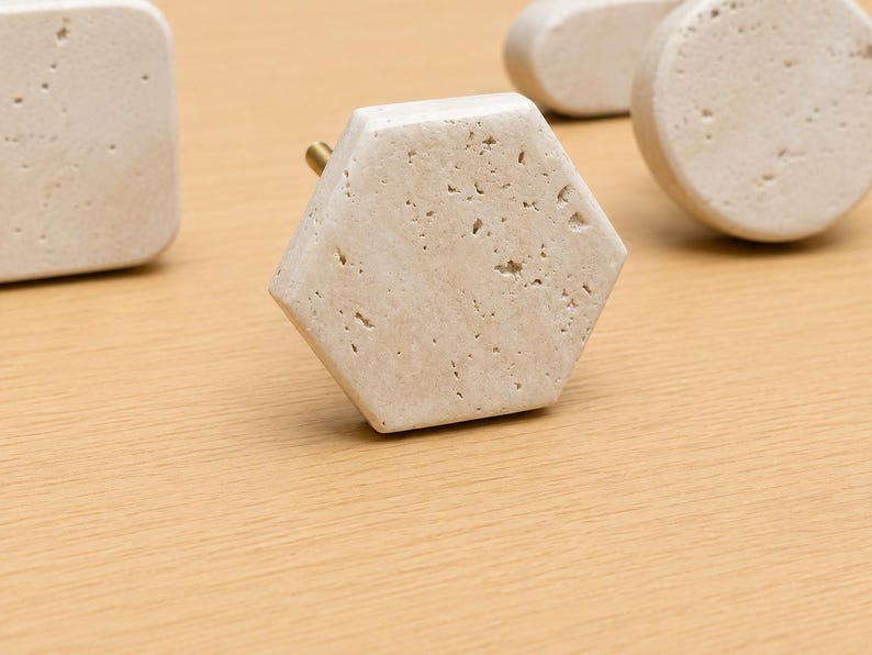 May include: A collection of cabinet knobs made from beige porous stone with natural pitting. The set includes a central hexagonal knob, a square knob to the left, and a circular knob to the right, each featuring a small brass-colored metal mounting post.