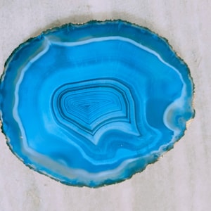 May include: A blue agate slice with a white rim and concentric rings of blue and white. The agate is polished and has a smooth surface.