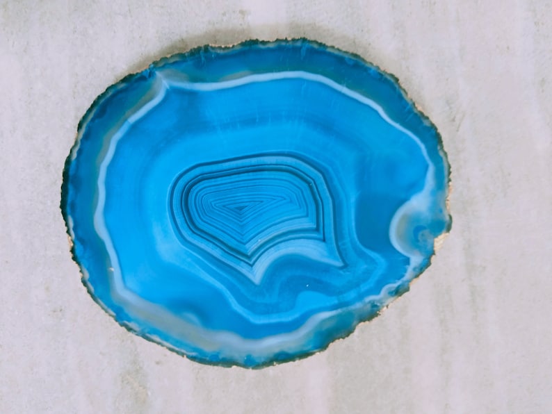 May include: A blue agate slice with a white rim and concentric rings of blue and white. The agate is polished and has a smooth surface.