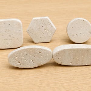May include: A collection of five light beige stone knobs featuring a porous, textured surface. The set includes one square, one hexagonal, one circular, and two elongated oval-shaped pieces, each with rounded edges and a natural, matte finish.