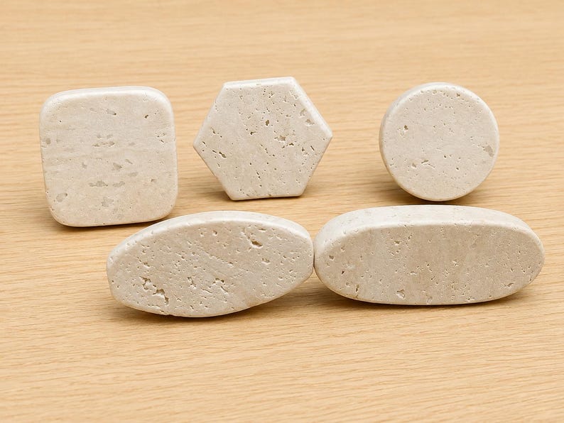 May include: A collection of five light beige stone knobs featuring a porous, textured surface. The set includes one square, one hexagonal, one circular, and two elongated oval-shaped pieces, each with rounded edges and a natural, matte finish.