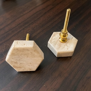May include: Two hexagonal cabinet knobs made of light beige stone with a natural speckled texture. Each knob features a central gold-toned metal bolt with a threaded shaft and a small nut attached to the hardware extending from the back.
