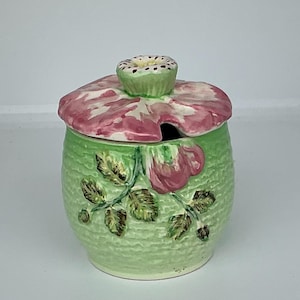 May include: A ceramic sugar bowl with a light green textured body featuring a raised pink rose and green leaf design. The lid is shaped like a pink flower petal with a small, white, dotted centre knob and a cutout for a spoon.