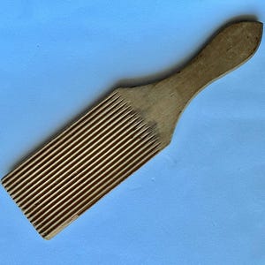 May include: A light brown wooden butter paddle featuring a rectangular head with a series of parallel, vertical grooves carved into the surface and a smooth, tapered handle extending from the top for a comfortable grip during use.