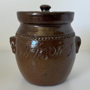 May include: A dark brown glazed ceramic crock with a matching lid and two side handles. The word Dripping is embossed in cursive script on the front, positioned below a decorative beaded band that encircles the upper portion of the vessel.