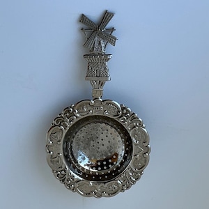 May include: A silver-coloured metal tea strainer featuring a circular bowl with a perforated mesh centre and an ornate rim decorated with embossed scenes. The handle is shaped like a detailed windmill structure with a rotating blade design.