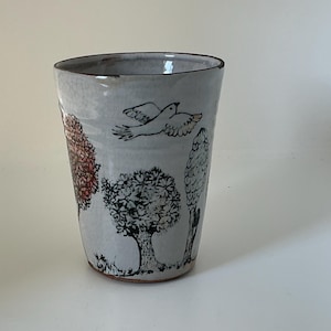 May include: A tapered ceramic tumbler with a crackled white glaze featuring black ink illustrations of various trees and a flying bird. The design includes a tree with reddish-orange foliage, a central leafy tree, and a tall slender tree, all set against a light background.