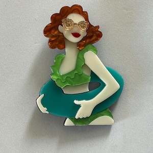 May include: An acrylic brooch depicting a figure with wavy auburn hair, wearing gold-toned glitter sunglasses and a bright green ruffled bikini top. The figure holds a large, solid teal-coloured circular swim ring around their waist.