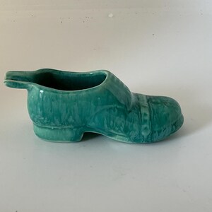 May include: A small, hollow ceramic shoe figurine with a glossy, mottled teal green glaze. The object features a low-cut opening at the top, a rounded toe, and a small tab extending from the heel area, all finished in a smooth, reflective surface.