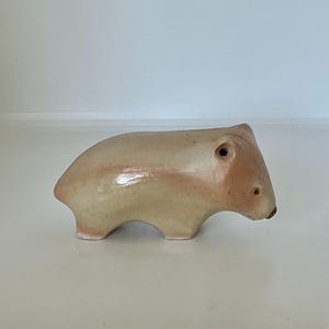 May include: A smooth, beige ceramic figurine shaped like a small, stout animal with a rounded back, short legs, and a pointed snout. The surface has a glossy, glazed finish with subtle variations in colour, featuring a small dark eye and nose.