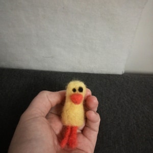 May include: A small needle-felted yellow duckling figurine with a fuzzy texture. The duckling features two small black circular eyes, a bright orange beak, and two orange feet extending from the bottom of its rounded yellow body.