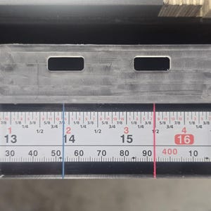 May include: A clear plastic ruler with two rectangular cutouts, positioned over a white and black measuring tape. The tape displays inch and centimetre markings, with numbers from 13 to 17. Vertical blue and red lines are also visible.