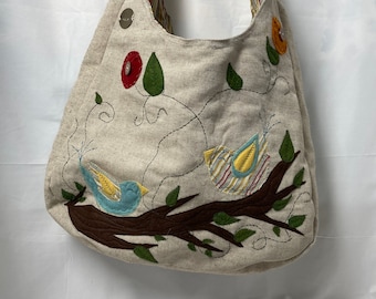 May include: Beige shoulder bag with embroidered birds and floral accents. The bag features a brown branch design with two birds, green leaves, and colorful flowers. The strap has a striped pattern. The bag is made of a textured fabric.