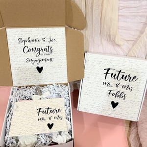 May include: Two white gift boxes with black lettering. The top box has a handwritten pattern and says "Stephanie & Joe. Congrats on your Engagement". The bottom box says "Future Mr. & Mrs. Fobbs".