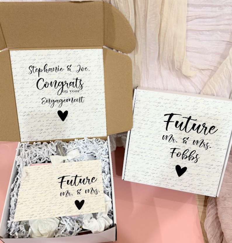 May include: Two white gift boxes with black lettering. The top box has a handwritten pattern and says "Stephanie & Joe. Congrats on your Engagement". The bottom box says "Future Mr. & Mrs. Fobbs".