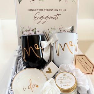 A gift box filled with wedding-themed items, including two black and white mugs with the words "Mr." and "Mrs." written in gold script, a small white candle with the words "Love & Laughter & Happily Ever After" written in gold script, a white rose, and a white dish with the words "I do" written in gold script.