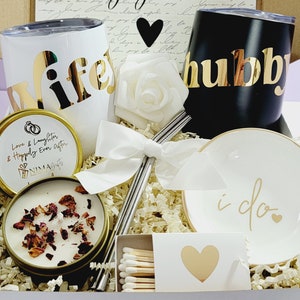 May include: A gift box filled with wedding-themed items, including two tumblers with the words "wife" and "hubby" in gold lettering, a candle with the text "Love & Laughter & Happily Ever After", a small white dish with the words "i do" in gold lettering, a white rose, and a box of matches.