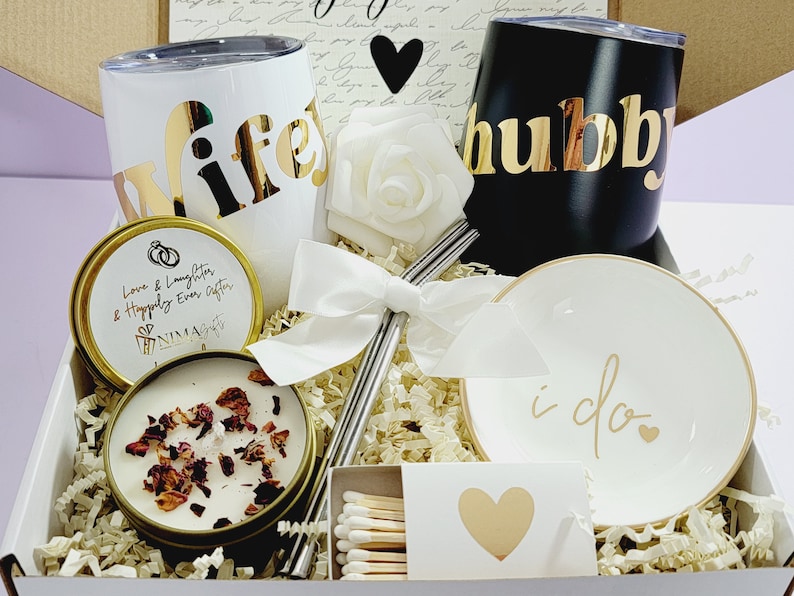 May include: A gift box filled with wedding-themed items, including two tumblers with the words "wife" and "hubby" in gold lettering, a candle with the text "Love & Laughter & Happily Ever After", a small white dish with the words "i do" in gold lettering, a white rose, and a box of matches.