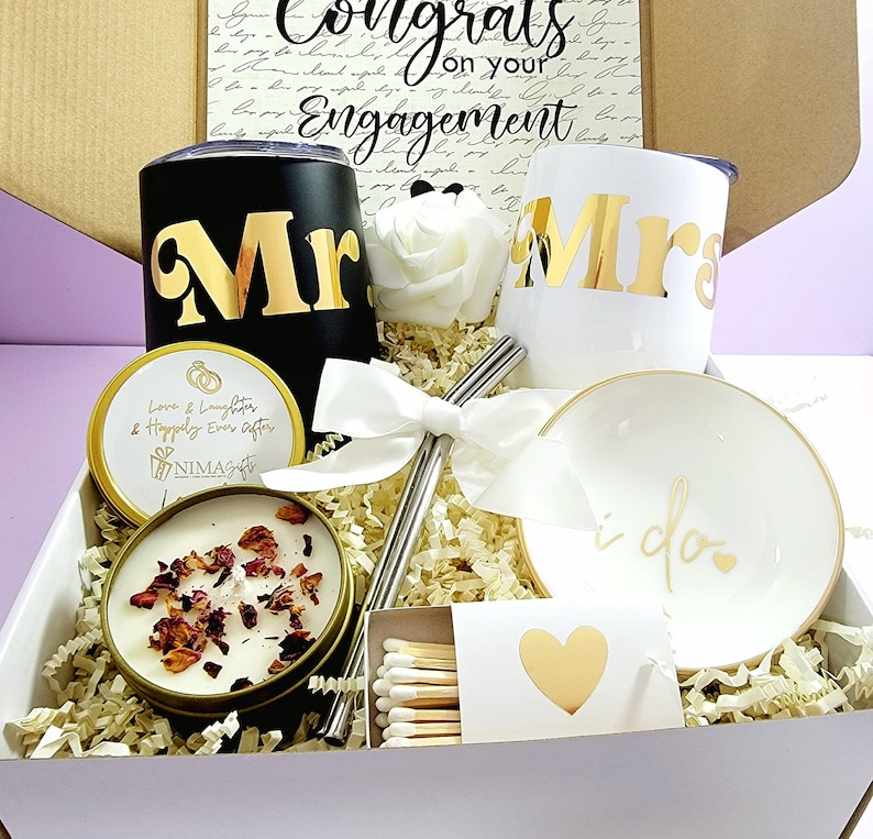 May include: A white gift box filled with engagement gifts. The box contains two black and white tumblers with gold lettering that says "Mr." and "Mrs.", a candle with rose petals, a small white dish with gold lettering that says "i do", two silver straws, a box of matches, and a white bow.