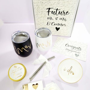 May include: A wedding gift set with a white box with the text "Future Mr. & Mrs. O'Connor" on it, two black and white tumblers, a white candle with the text "Love & Laughter Happily Ever After IMA of wonder", a white ring dish with the text "i do", a box of matches with a gold heart, a white rose, and a card with the text "Congrats on your Engagement!"