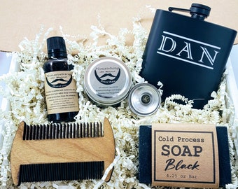 May include: A gift box filled with grooming products for men, including beard oil, beard balm, a wooden comb, a metal flask engraved with the name "DAN", and a bar of black soap. The box is filled with shredded paper.