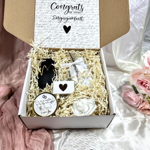 May include: A white gift box filled with engagement gifts. The box is open and the contents include a candle, a small card, a glass bottle, and a white rose. The box is decorated with a handwritten note that says "Alex & Caroline, Congrats on your Engagement".