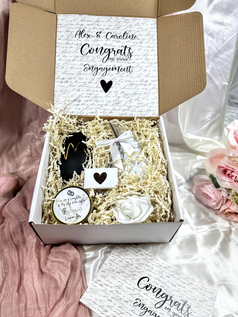 May include: A white gift box filled with engagement gifts. The box is open and the contents include a candle, a small card, a glass bottle, and a white rose. The box is decorated with a handwritten note that says "Alex & Caroline, Congrats on your Engagement".