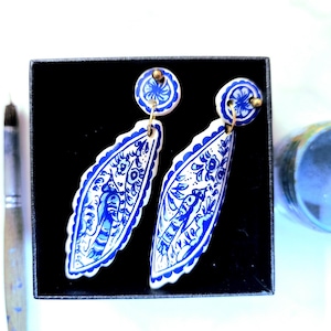 May include: A pair of white and blue ceramic earrings with a floral and bird design. The earrings are in a black box, with a paintbrush and a small container visible in the background. The earrings have a gold-colored stud.