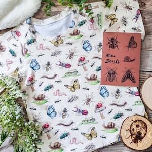 以下が含まれることがあります： 様々な昆虫、蝶、その他の小さな生き物のカラフルなプリントが施された白いベビーロンパース。ロンパースには「ミニビースト」という文字が印刷されています。