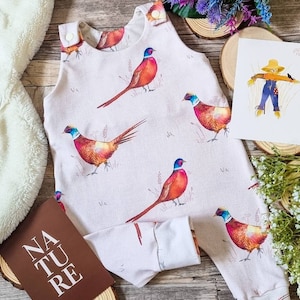 May include: A white baby onesie with a brown, white and red pheasant print. The onesie has brown trim around the neck and armholes. The onesie has the word "NATURE" printed on the front.