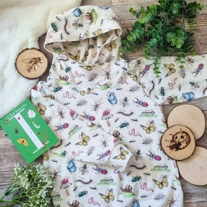 以下が含まれることがあります： カラフルな昆虫柄の白いフード付きスウェットシャツ。プリントには、蝶、甲虫、アリなど、さまざまな昆虫が描かれています。スウェットシャツには、緑色のアルファベットカードが付いており、'I'と'I is for insects'という文字が書かれています。