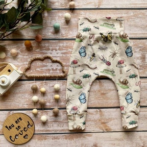 May include: Beige baby pants with a print of various insects, butterflies, and mushrooms. The pants have a wide elastic waistband and cuffed ankles.
