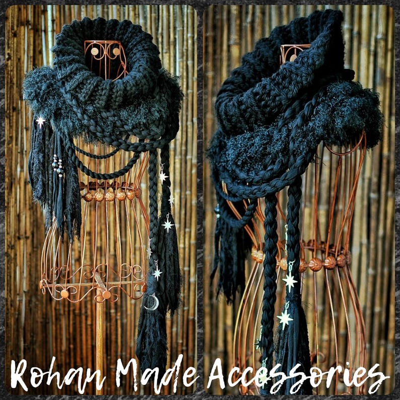 May include: A black chunky knit infinity scarf featuring a thick ribbed collar, layered braided cords, and dangling fringe. The scarf is adorned with silver-toned star and crescent moon charms. The text Rohan Made Accessories is visible.