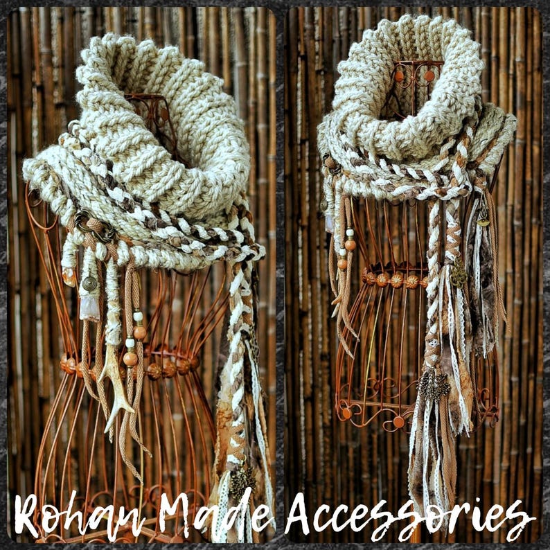 May include: A chunky knit beige infinity scarf featuring a thick ribbed texture, adorned with dangling braided fabric strips, wooden beads, and small metallic charms. The scarf is displayed on a wire mannequin form with the text Rohan Made Accessories.