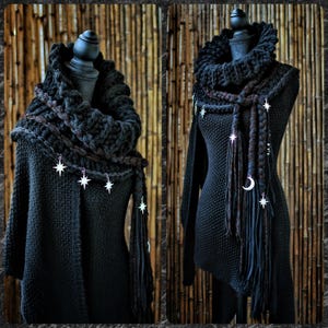 May include: A black knitted cowl scarf featuring a thick, chunky texture and draped around a mannequin. The scarf is adorned with silver-toned metal charms shaped like stars and a crescent moon, attached to long, braided fabric fringe accents.