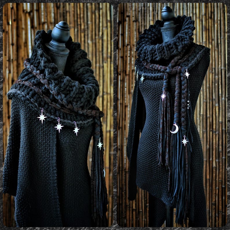May include: A black knitted cowl scarf featuring a thick, chunky texture and draped around a mannequin. The scarf is adorned with silver-toned metal charms shaped like stars and a crescent moon, attached to long, braided fabric fringe accents.