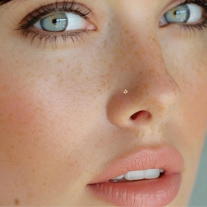 May include: Close-up of a person's face with a small gold nose ring. The person has freckles and light skin.