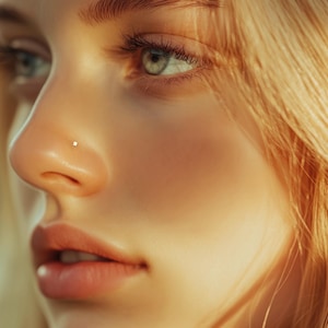 May include: Close-up of a woman's face with blonde hair. She has a small nose piercing and her eyes are looking down and to the left. The light is hitting her face from the right, creating a soft glow.