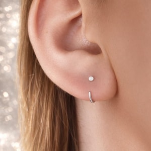 May include: Close-up of an ear with two silver earrings. One is a small stud, and the other is a small hoop earring. The ear is set against a blurred background of blonde hair.