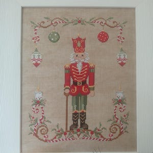Può includere: Opera d'arte a punto croce di un soldato schiaccianoci natalizio in uniforme rossa, pantaloni verdi e stivali neri. Il pezzo incorniciato presenta ghirlande festive, ornamenti e candele. Lo sfondo è di colore beige neutro.