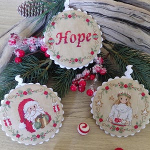 Può includere: Tre ornamenti natalizi rotondi e ricamati con bordi smerlati. Un ornamento recita "HOPE" a punto croce rosso. Gli altri due raffigurano Babbo Natale e un angelo. Gli ornamenti sono decorati con motivi floreali rossi e verdi.