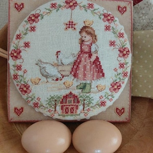 Può includere: Ricamo a punto croce raffigurante una bambina con galline, un fienile e dettagli floreali. Il ricamo è incorniciato in una cornice quadrata con dettagli a cuore. Due uova marroni sono in una ciotola di legno, completando il tema pastorale.