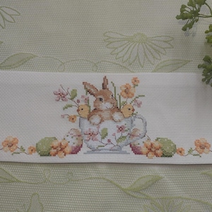 Peut inclure: Une broderie au point de croix blanche représentant un lapin et deux poussins dans une tasse à thé, entourés de fleurs et d'œufs de Pâques. Le motif est bordé d'éléments floraux, créant un thème printanier ou pascal.