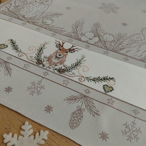 May include: A beige and brown embroidered table runner with a winter theme. The design includes a deer, pine cones, snowflakes, and an owl. The runner is made of a soft fabric and has a decorative border.