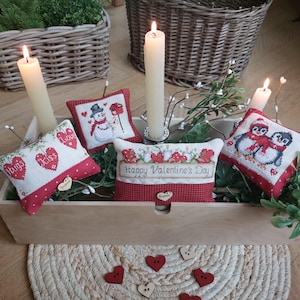 Può includere: Quattro piccoli cuscini ricamati in rosso e bianco con temi di San Valentino. I cuscini sono disposti in una scatola di legno con un tappetino di paglia intrecciato sotto. I cuscini presentano un pupazzo di neve, pinguini, cuori e il testo "Happy Valentine's Day".