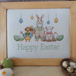 Può includere: Un ricamo a punto croce con un coniglio, un agnello e un pulcino con il testo "Buona Pasqua". Il disegno è incorniciato in una cornice di legno chiaro.