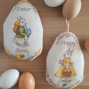 Pode incluir: Duas almofadas bordadas em forma de ovo de Páscoa com desenhos de coelhos. Uma almofada apresenta um coelho segurando um ovo e diz "Easter Bunny". A outra almofada apresenta um coelho segurando uma cesta e diz "Happy Easter".