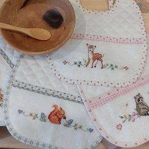 May include: Four white baby bibs with embroidered woodland animal designs. The bibs have a pocket on the bottom and a pink, blue, or white trim.
